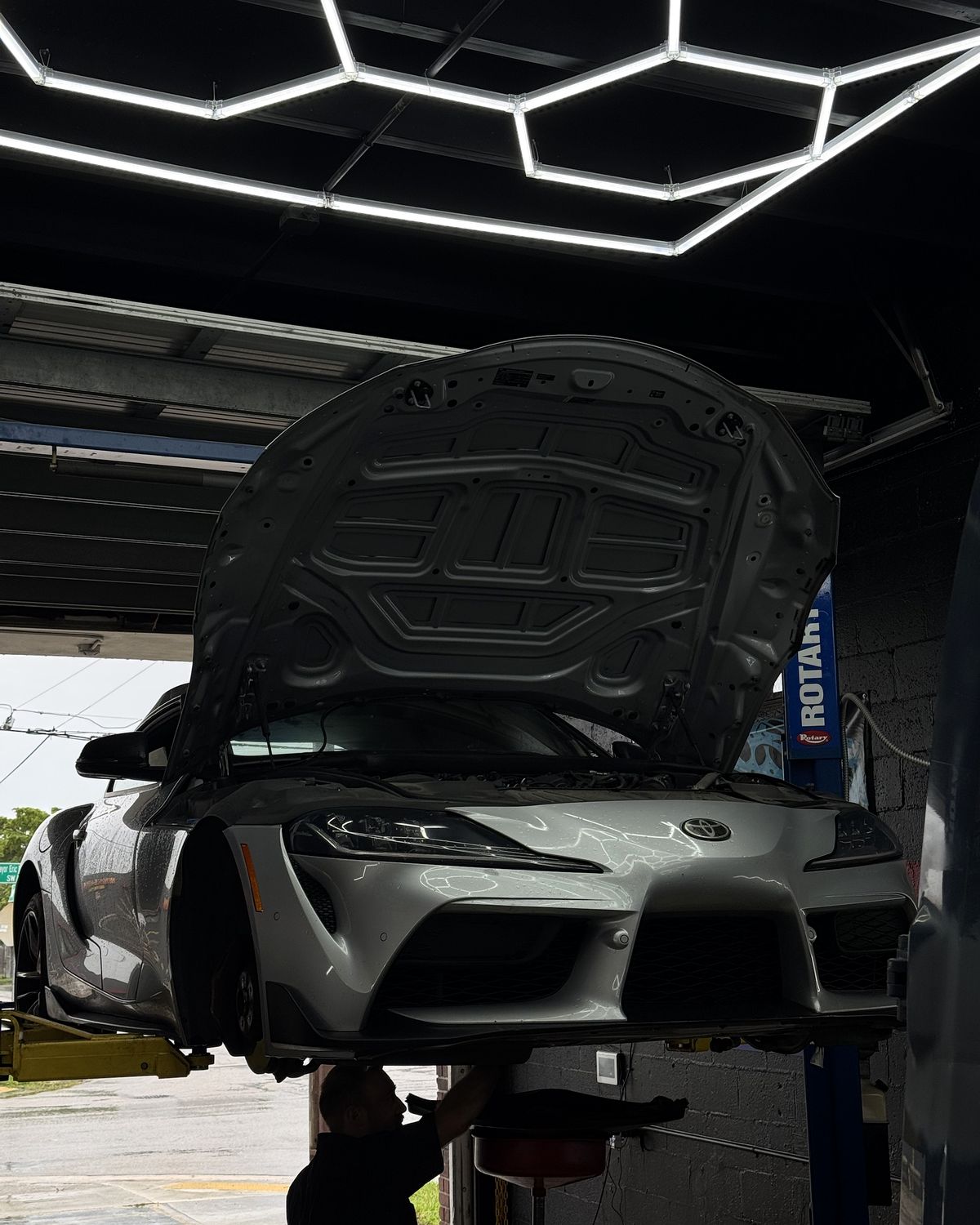 Silver Toyota GR Supra on the lift with the hood up while a technician drains the oil