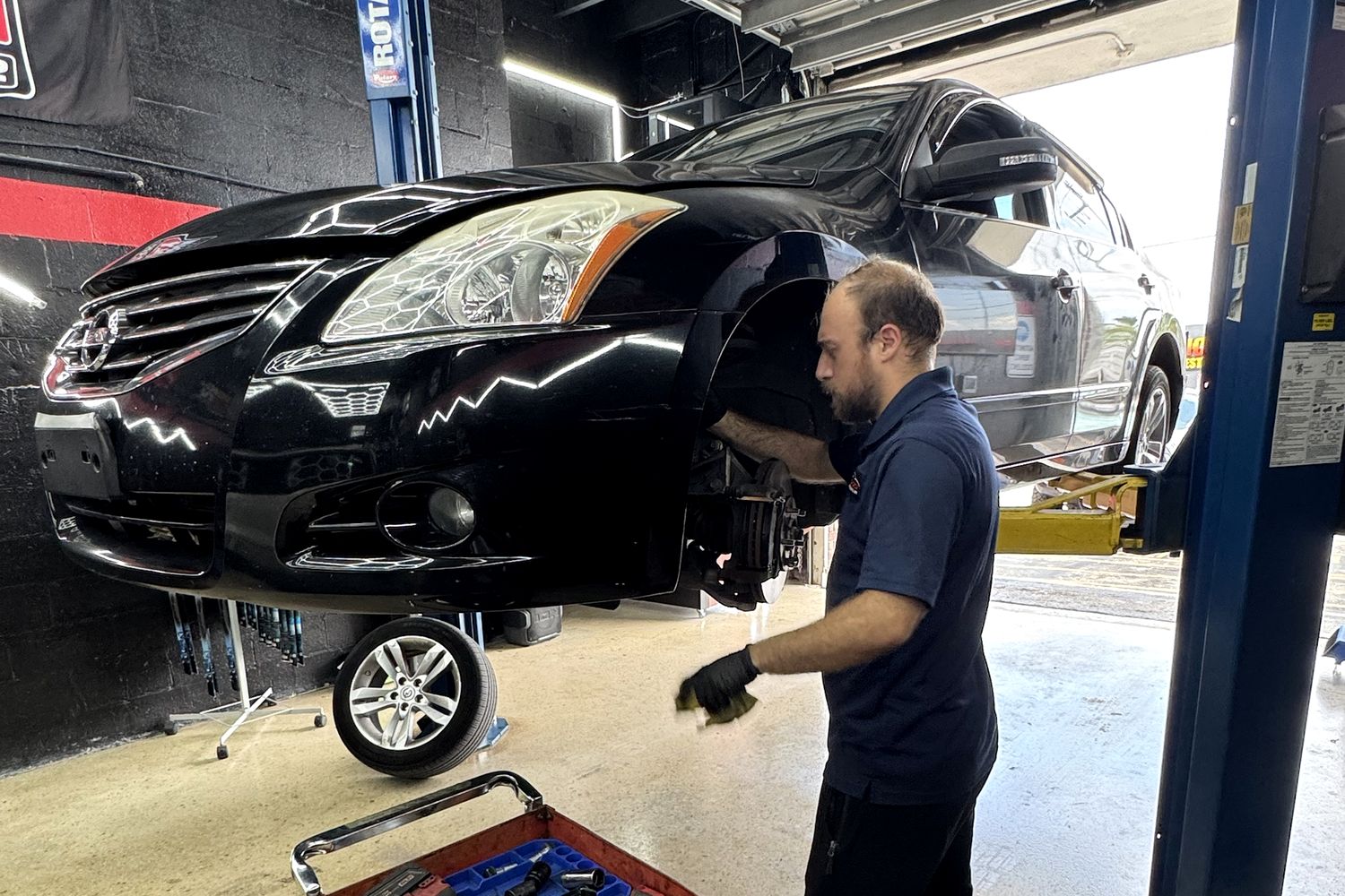 Technician replacing the front brakes on a black Nissan Altima, new rotor and pads on the tool cart